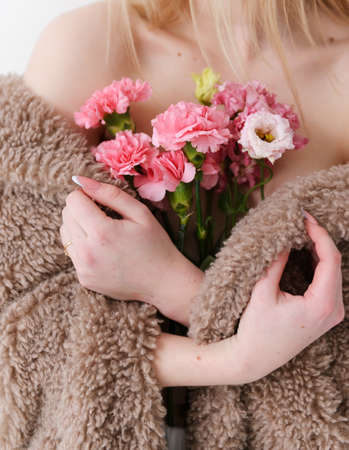 beautiful white young blonde woman in brown furry coat holds a bouquet of pink flowers in her arms closely on a white backgroundの写真素材