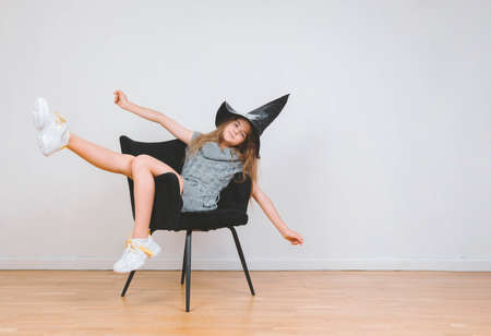 White little girl sits on a chair in witch hat halloween freely in grey dress full-lengthの写真素材
