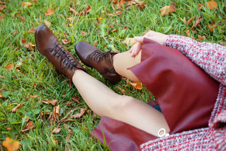 Young woman's legs sitting in brown boots and in burgundy skirt on grass with autumn leaves horizontal texture nature fall october sexyの写真素材