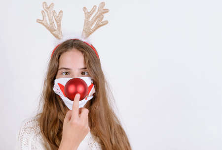 Young girl in white sweater, musk and horns touches red bouble as nose christmas simple background minimalistic holidaysの写真素材