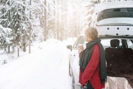 white beautiful blonde young woman in a red sweater, trunk of a car in the woods winter road among...