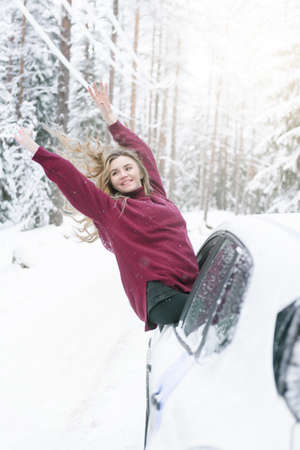 1 white young blonde woman in a red sweater leaned out of the car window on a winter road in the woods, smilesの写真素材