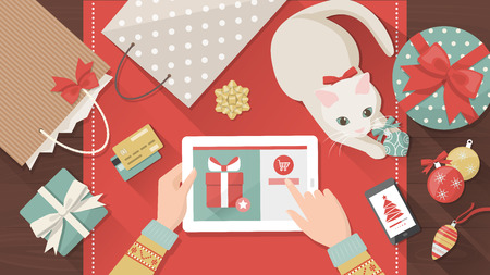 Woman purchasing Christmas gifts online using a tablet, her cat is playing with a bauble on the desk, holiday and celebrations bannerのイラスト素材
