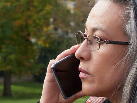 Older woman with gray hair and glasses wearing and talking on a mobile phone. Sitting on stone...