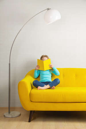 a blonde girl is sitting on the sofa in the room and reading a book in a yellow cover