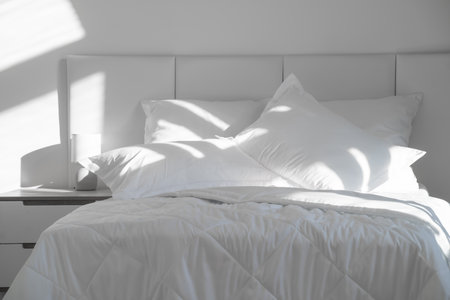A bed with white pillows and a blanket, next to a bedside table with a lamp, daylightの写真素材