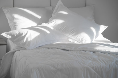 A bed with white pillows and a blanket, next to a bedside table with a lamp, daylightの写真素材