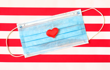 Disposable medical mask on striped red white background with red heart on it against bacteria and...