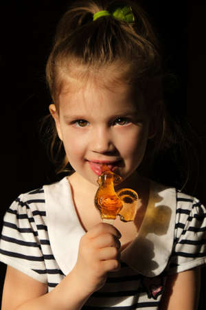 Little girl with lollypop in hand, Soft sunset light and shadow. Cute child licks delicious candy, in dark settings.の写真素材