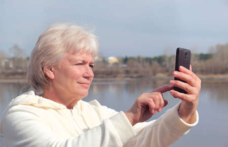 Portrait of happy blonde senior woman holding camera and taking selfie. Selective focus, Focus on...
