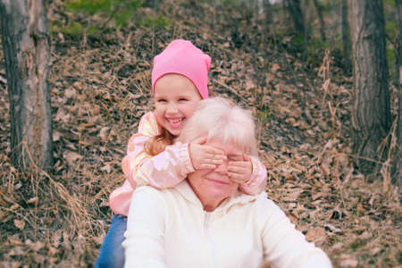 Granddaughter closing grandmother eyes with hands, playing funny game outdoors, family having fun togetherの写真素材
