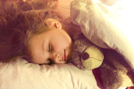 Sleeping little girl in pjamas with teddy bear lie down on bed while sun shine through the window,...