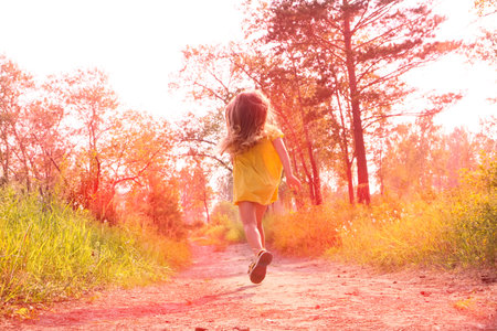 Happy little girl with blond long hair in a yellow dress and running along path on meadow with sunsetの写真素材