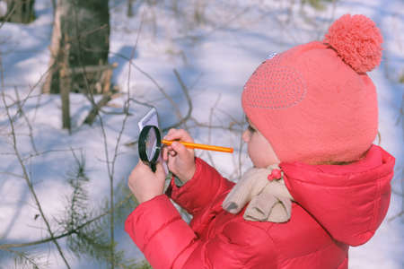 Little girl in warm winter clothes with a magnifying glass in her hand investigate details of nature. Winter outdoor kids activity and learning conceptの写真素材