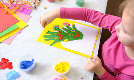 Little girl making homemade greeting card from envelope, paper own palms and decoration clay,...