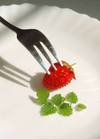 single raw ripe strawberry on a white plate with mint leaves and a forkの写真素材