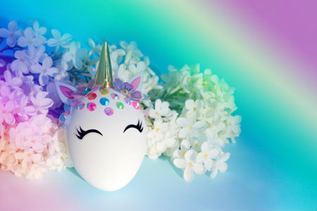 Creative decorative Easter egg in the form of a unicorn with white delicate flowers around on a rainbow backgroundの写真素材