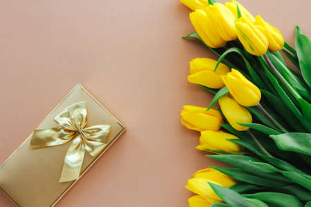 yellow tulips and a gift box with a gold bow on a beige background. Mother Day background. Beautiful...