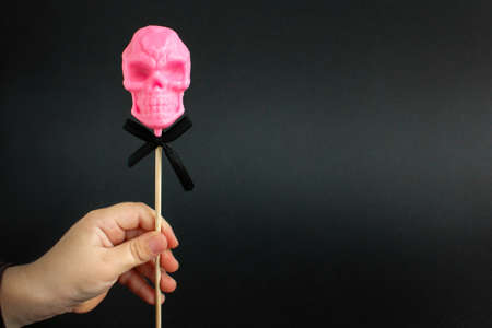 one pink lollipop in the shape of a skull on a stick in a childs hand on a black background. halloween concept.の写真素材