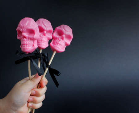 three pink lollipops in the shape of a skull on a stick in a child's hand on a black background. halloween concept, trick or treatの写真素材