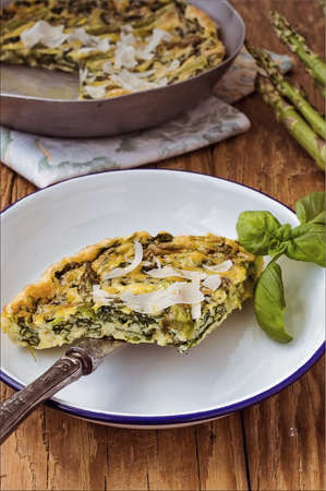 Italian omelette omelet slices on a white enamel plate with a knife accompanied by a frying pan with cooked omelette and fresh asparagusの写真素材