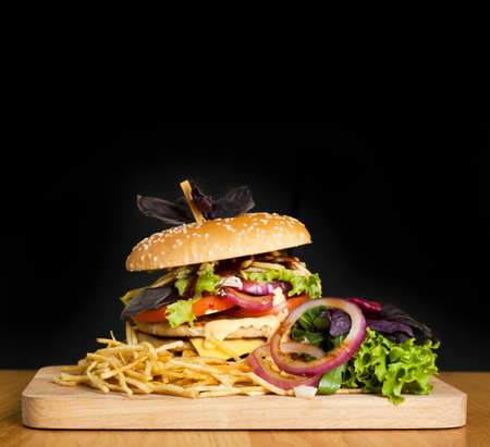 A delicious burger with two cutlets, basil, thin fries, lettuce and onion rings on a wooden board on the dark background. Photo causing appetite. The concept of fast food, delicious but unwholesome food.の写真素材