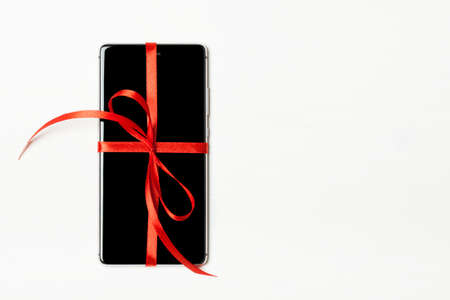 Phone as a gift for your loved one. Smart phone with a red bow on a light wooden background. The concept of the day of St. Valentine, love, surprises. Flat flyの写真素材