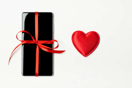 Phone as a gift for your loved one. Smart phone with a red bow on a light wooden background with heart. The concept of the day of St. Valentine, love, surprises. Flat flyの写真素材
