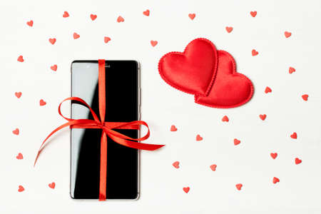 Phone as a gift for your loved one. Smartphone with a red bow on a light wooden background decorated with hearts. The concept of the day of St. Valentine, love, surprises. Flat flyの写真素材
