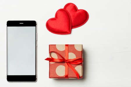 Phone and a gift for your loved one. Smartphone with a red bow and gift on a light wooden background decorated with hearts. The concept of the day of St. Valentine's, weddings, engagements, Mother's Day, birthday, New Year, Christmas, holidays. Flat flyの写真素材