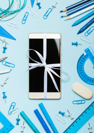 School supplies of blue and white colors on a blue background and new phone as a gift. Male or...