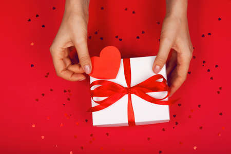 Beautiful female hand with minimalistic gift on a red background with cute heart. Concept of the...