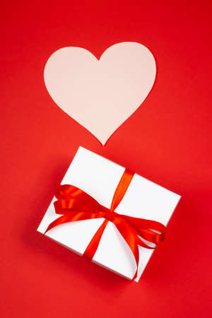 A gift with paper for notes in the shape of a heart on a red background. Surprise your loved one. The concept of the St. Valentines day, birthday and other holidays. Flat layの写真素材