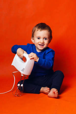 A pretty toddler boy with a mixer. Cute mothers assistant. Trending colors of clothing and...