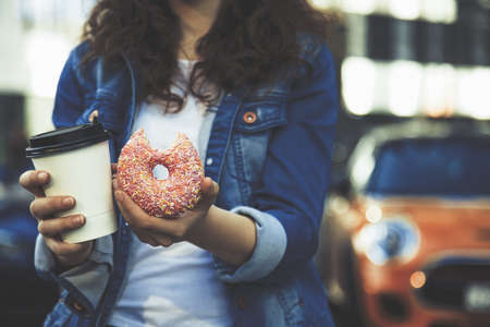 hands holding a cup of coffee and pink donut in streetの写真素材