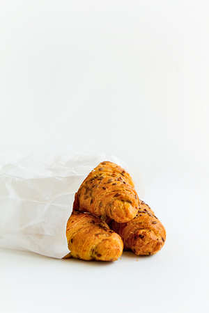 three delicious croissants with seeds on the baking surface in a white paper bag on a white table,...