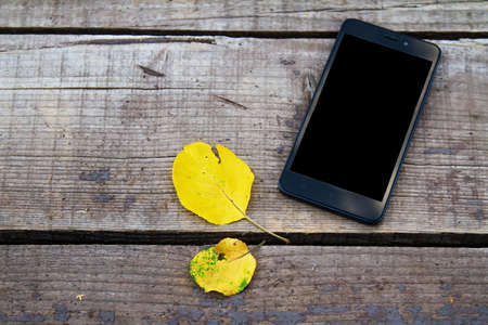 black mobile phone on a wooden old autumn background, near the yellow autumn leaves.Copy spaceの写真素材