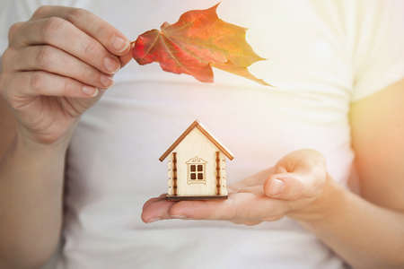 Small wooden house in a woman's hand. The second hand holds autumn maple red leaf, protecting roof down homes from stormy weather and sun. Close-up, the concept of protecting your homeの写真素材