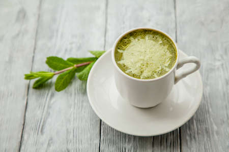 white cup of hot matcha green tea latte and branch of mint on light wooden background
