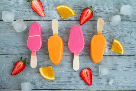 four pink and yellow ice creams on a stick on a gray wooden background next to juicy pieces of fruit and ice.の写真素材