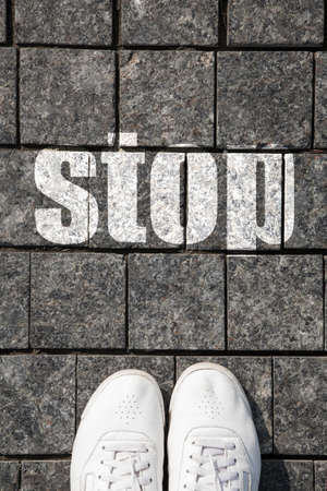 Feet with white sneakers, standing on the cobblestone street of the city with a written stop sign. Concept.の写真素材