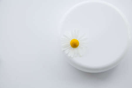 White jar of moisturizer and chamomile flowers on a white background.