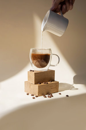 Coffee in a glass cup on a beige background. The hand pours milk into it. Minimalist stylish photography in daylight with shadows and light.の写真素材