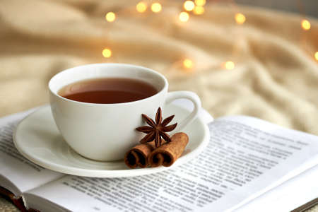 White cup of tea with cinnamon on a n open book and beige plaid. Christmas lights.の写真素材