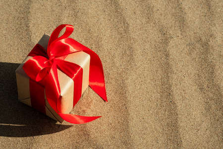 Gift in craft paper with satin ribbon on a sandy background. Celebrating Christmas on vacation