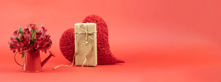 Red alstroemerias in a watering can with a gift in craft paper and red heart on St. Valentines Day. Bannerの写真素材