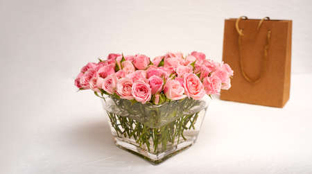 Bouquet of pink roses in a glass vase with a gift in a craft bag on a light backgroundの写真素材