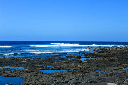 black rocky beach with small waves and clear blue skyの写真素材