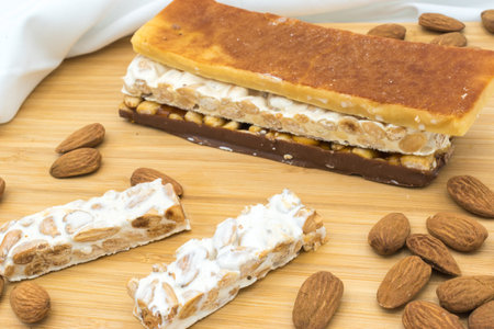 Different types of turrÃ³n (nougat), a traditional Christmas sweet in Spain on a wooden surface, surrounded by almonds and a white tablecloth.の写真素材