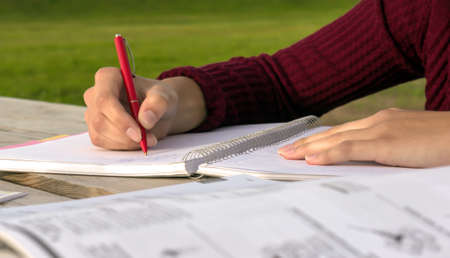 outdoors at a wooden table female hand writing in a notebook, burgundy sweater, green grass in the background, the book lying next, closeupの写真素材